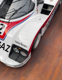 A vintage race car with a sleek aerodynamic design featuring a white body with red and blue accents. The car has prominent branding and sponsor logos, including 'futurhome' and 'Bilstein'. It is equipped with two large headlamps and sits on a wooden floor.