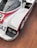 A vintage race car with a sleek aerodynamic design featuring a white body with red and blue accents. The car has prominent branding and sponsor logos, including 'futurhome' and 'Bilstein'. It is equipped with two large headlamps and sits on a wooden floor.