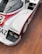 A vintage race car with a sleek aerodynamic design featuring a white body with red and blue accents. The car has prominent branding and sponsor logos, including 'futurhome' and 'Bilstein'. It is equipped with two large headlamps and sits on a wooden floor.