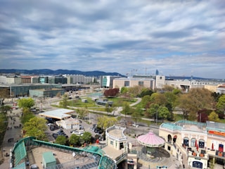 A panoramic view of an urban landscape featuring modern buildings, construction cranes, and a park with green spaces. In the foreground, a small amusement area with a carousel and people walking along pathways. The background showcases a hilly terrain and a cloudy sky.