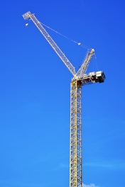 A tall yellow construction crane with a lattice boom structure stands against a clear blue sky. The crane's arm extends diagonally upwards to the left, and there is a small cabin attached near the top of the structure.