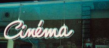 A retro-style neon sign displaying the word 'Cinéma' is hanging against a window, with faint reflections of the surrounding city architecture visible in the glass. Behind the sign, there are partially obscured black-and-white movie posters, enhancing the vintage atmosphere.