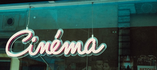 A retro-style neon sign displaying the word 'Cin&eacute;ma' is hanging against a window, with faint reflections of the surrounding city architecture visible in the glass. Behind the sign, there are partially obscured black-and-white movie posters, enhancing the vintage atmosphere.