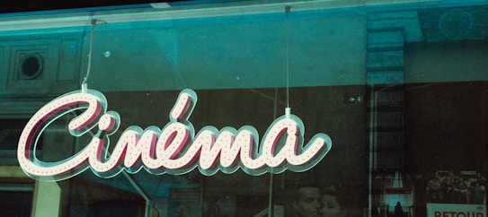 A retro-style neon sign displaying the word 'Cinéma' is hanging against a window, with faint reflections of the surrounding city architecture visible in the glass. Behind the sign, there are partially obscured black-and-white movie posters, enhancing the vintage atmosphere.