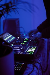 A DJ setup with turntables, mixers, and glowing LED lights in a dimly lit environment. A person is operating the equipment, adjusting the controls. The overall ambiance is cool with blue lighting, adding to a modern and energetic atmosphere.