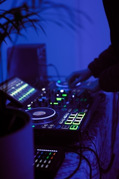 A DJ setup with turntables, mixers, and glowing LED lights in a dimly lit environment. A person is operating the equipment, adjusting the controls. The overall ambiance is cool with blue lighting, adding to a modern and energetic atmosphere.