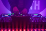 A DJ booth with colorful lights and equipment, including a Denon DJ controller. A person is adjusting the controls, creating a vibrant and dynamic atmosphere with purple and blue hues.