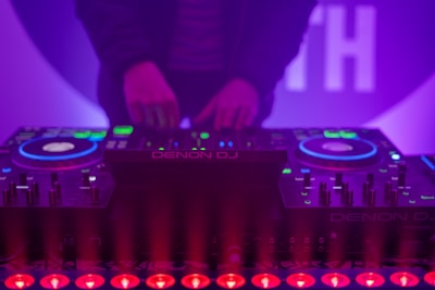 A DJ booth with colorful lights and equipment, including a Denon DJ controller. A person is adjusting the controls, creating a vibrant and dynamic atmosphere with purple and blue hues.