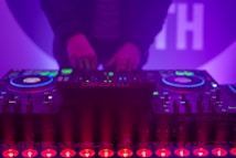 A DJ booth with colorful lights and equipment, including a Denon DJ controller. A person is adjusting the controls, creating a vibrant and dynamic atmosphere with purple and blue hues.