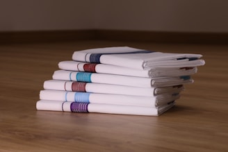 A neat stack of six white folded handkerchiefs is placed on a wooden surface. Each handkerchief features a distinct stripe of color along one edge, including shades of blue, red, and purple. The arrangement and lighting highlight the crispness and texture of the fabric.