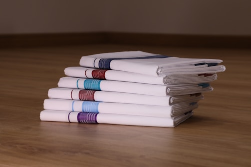 A neat stack of six white folded handkerchiefs is placed on a wooden surface. Each handkerchief features a distinct stripe of color along one edge, including shades of blue, red, and purple. The arrangement and lighting highlight the crispness and texture of the fabric.