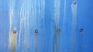 A textured blue metal surface with vertical rust stains and multiple rivets evenly spaced across it.