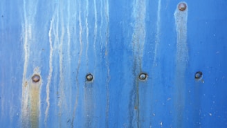 A textured blue metal surface with vertical rust stains and multiple rivets evenly spaced across it.