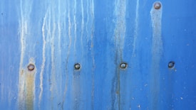 A textured blue metal surface with vertical rust stains and multiple rivets evenly spaced across it.