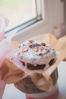 A beautifully decorated cake with white frosting, topped with chopped nuts and small pieces of rose petals. It is wrapped in brown parchment paper, placed near a window, allowing natural light to highlight the textures and colors.