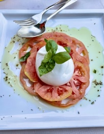 A fresh salad dish consisting of sliced tomatoes arranged in a circular pattern with a ball of mozzarella cheese placed on top. Fresh basil leaves are placed on the cheese, and olive oil drizzles surround the arrangement on a white rectangular plate. A fork and spoon are placed at the top side of the plate.