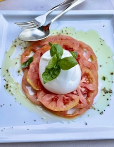 A fresh salad dish consisting of sliced tomatoes arranged in a circular pattern with a ball of mozzarella cheese placed on top. Fresh basil leaves are placed on the cheese, and olive oil drizzles surround the arrangement on a white rectangular plate. A fork and spoon are placed at the top side of the plate.