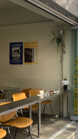 A minimalist cafe interior with sleek metal tables and light brown chairs. On the wall, there are two posters related to architecture, one in blue and another in yellow. In the corner, a hanging plant adds a touch of greenery. The table is set with a white cup, a few plates, and some cutlery. The setting is bathed in natural light coming from the large glass window on the right.