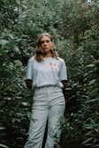 A person stands amidst dense green foliage, wearing a light-colored shirt with 'GP & F' printed on it. Their expression is neutral, and the lush greenery surrounds them, creating a natural, immersive setting.