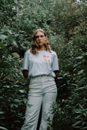 A person stands amidst dense green foliage, wearing a light-colored shirt with 'GP & F' printed on it. Their expression is neutral, and the lush greenery surrounds them, creating a natural, immersive setting.