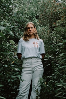 A person stands amidst dense green foliage, wearing a light-colored shirt with 'GP & F' printed on it. Their expression is neutral, and the lush greenery surrounds them, creating a natural, immersive setting.