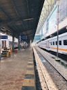 A train station platform with a railway track on the right, lined by stationary trains under a metal canopy. Signage displays directions and destinations, while lighting fixtures are suspended from the ceiling. The station is calm, showing a clear view down the length of the platform.