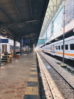 A train station platform with a railway track on the right, lined by stationary trains under a metal canopy. Signage displays directions and destinations, while lighting fixtures are suspended from the ceiling. The station is calm, showing a clear view down the length of the platform.