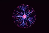 A glowing plasma ball emits vibrant pink and blue filaments of light resembling electrical discharges. These filaments radiate from a central orb and create an intricate web-like pattern against a dark background.