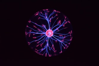 A glowing plasma ball emits vibrant pink and blue filaments of light resembling electrical discharges. These filaments radiate from a central orb and create an intricate web-like pattern against a dark background.