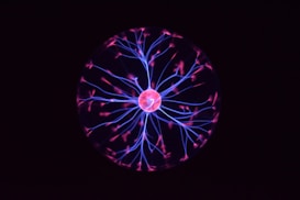 A glowing plasma ball emits vibrant pink and blue filaments of light resembling electrical discharges. These filaments radiate from a central orb and create an intricate web-like pattern against a dark background.