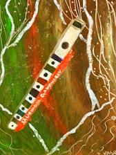 A painting features a wooden flute with multiple holes resting diagonally against an abstract background. The flute has a visible label with text in red. Surrounding it are swirling white lines and splashes of green, brown, and red, creating a dynamic and lively composition.