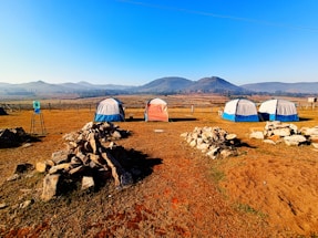 A serene campsite features five tents arranged on a dry grass field, surrounded by rocky piles. Beyond the tents, rolling hills stretch into the distance under a clear blue sky. A water cooler is placed on a stand to the left, adding a practical element to the tranquil outdoor setting.