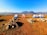 A serene campsite features five tents arranged on a dry grass field, surrounded by rocky piles. Beyond the tents, rolling hills stretch into the distance under a clear blue sky. A water cooler is placed on a stand to the left, adding a practical element to the tranquil outdoor setting.