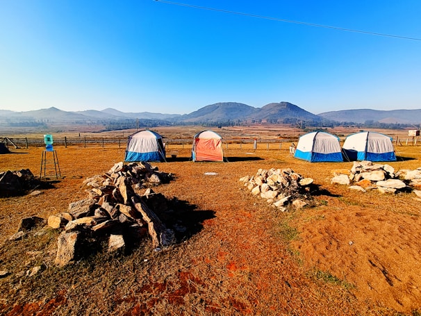 A serene campsite features five tents arranged on a dry grass field, surrounded by rocky piles. Beyond the tents, rolling hills stretch into the distance under a clear blue sky. A water cooler is placed on a stand to the left, adding a practical element to the tranquil outdoor setting.