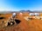 A serene campsite features five tents arranged on a dry grass field, surrounded by rocky piles. Beyond the tents, rolling hills stretch into the distance under a clear blue sky. A water cooler is placed on a stand to the left, adding a practical element to the tranquil outdoor setting.