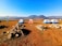 A serene campsite features five tents arranged on a dry grass field, surrounded by rocky piles. Beyond the tents, rolling hills stretch into the distance under a clear blue sky. A water cooler is placed on a stand to the left, adding a practical element to the tranquil outdoor setting.