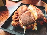 A close-up of a crispy fried chicken sandwich with a golden bun, served in a dark tray. The sandwich is filled with fried chicken pieces, lettuce, and sauce, placed on a wooden table with a warm, inviting ambiance.