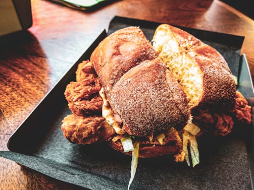 A close-up of a crispy fried chicken sandwich with a golden bun, served in a dark tray. The sandwich is filled with fried chicken pieces, lettuce, and sauce, placed on a wooden table with a warm, inviting ambiance.