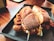 A close-up of a crispy fried chicken sandwich with a golden bun, served in a dark tray. The sandwich is filled with fried chicken pieces, lettuce, and sauce, placed on a wooden table with a warm, inviting ambiance.