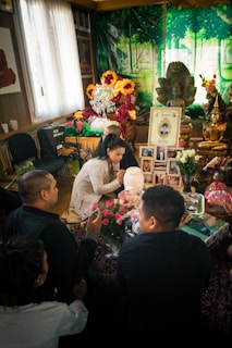 A group of people gathered in a room decorated with vibrant flowers, candles, and religious iconography, including a statue of Ganesh. Several framed photos and offerings are arranged on a table. The setting appears to be a shrine or a place of remembrance.
