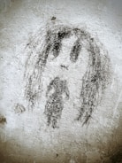 A rough black charcoal drawing on a textured wall depicts a simplistic human figure with long hair and a solemn expression. The artwork appears abstract and graffiti-like, occupying the central space with rough, uneven lines and shading.