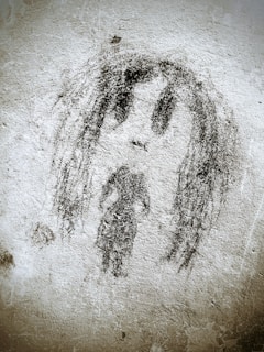A rough black charcoal drawing on a textured wall depicts a simplistic human figure with long hair and a solemn expression. The artwork appears abstract and graffiti-like, occupying the central space with rough, uneven lines and shading.