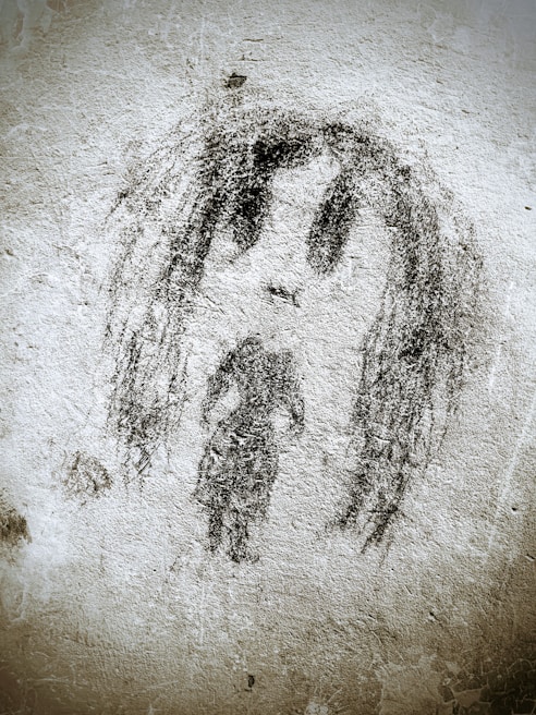 A rough black charcoal drawing on a textured wall depicts a simplistic human figure with long hair and a solemn expression. The artwork appears abstract and graffiti-like, occupying the central space with rough, uneven lines and shading.