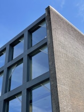 A modern building façade with a combination of large glass panels and dark structural framing. The structure features a brick wall on one side, adding a contrasting texture to the sleek glass and metal design. Clear blue skies are visible in the reflection of the glass windows.