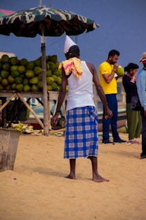 A man wearing a white tank top and a blue checked lungi stands on a sandy beach, facing away from the camera. He wears a hat made from a newspaper and a towel drapes over one shoulder. There is a stall to his left under a camouflaged umbrella, stocked with green coconuts. In the background, another man in a yellow shirt is drinking from a coconut, while two more people stand nearby.