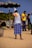 A man wearing a white tank top and a blue checked lungi stands on a sandy beach, facing away from the camera. He wears a hat made from a newspaper and a towel drapes over one shoulder. There is a stall to his left under a camouflaged umbrella, stocked with green coconuts. In the background, another man in a yellow shirt is drinking from a coconut, while two more people stand nearby.