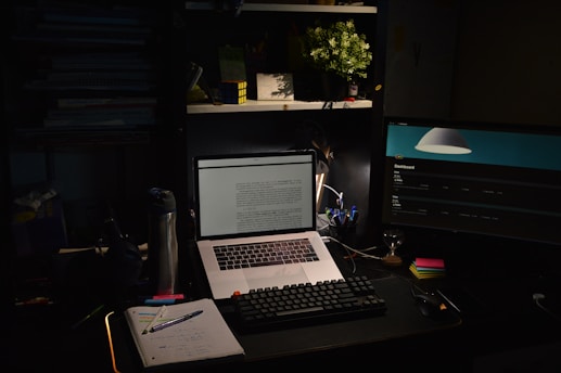 A dimly lit workspace featuring a laptop with text on the screen, a desktop monitor displaying a dashboard interface, a mechanical keyboard, and a notebook with writing utensils on a desk. There is a water bottle and an arrangement of sticky notes. Above the desk, shelves hold a potted plant and a Rubik's cube. The overall setup is minimalist yet functional, with a cozy ambience.