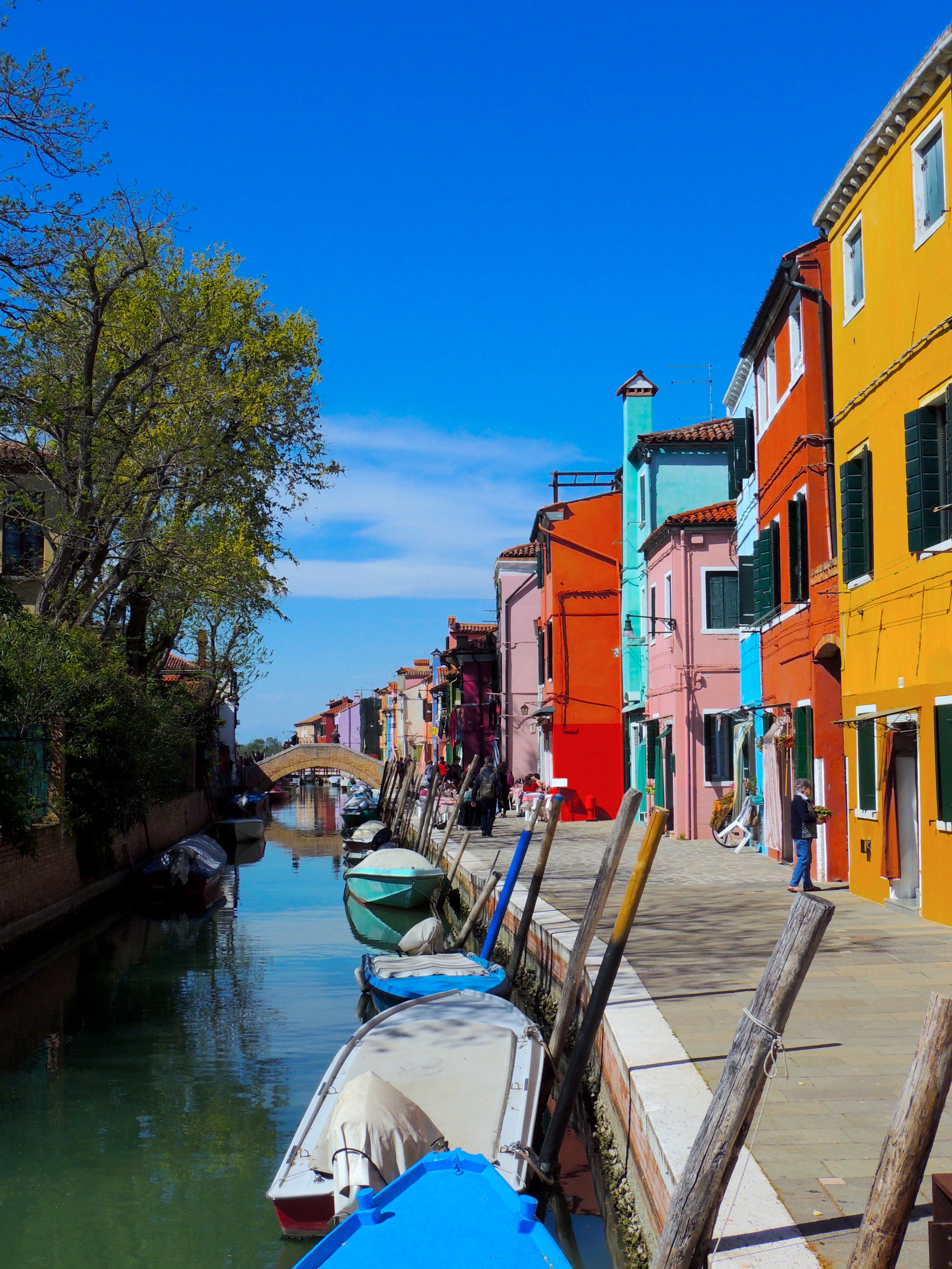 A canal on the island of Burano in the Venetian lagoon, lined with orange, pink and turquoise houses and small boats tied to mooring poles