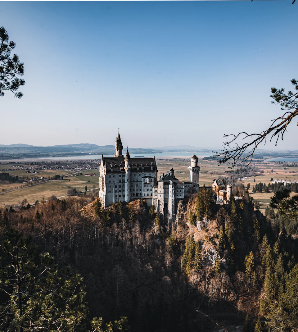 The image depicts a majestic castle perched atop a cliff, overlooking a valley below. The castle is constructed from stone and features multiple towers and turrets that reach towards the clear blue sky. The surrounding landscape includes lush green fields and trees, creating a picturesque backdrop for the castle's grandeur. The perspective of the photo is taken from a high vantage point, providing an expansive view of the castle and its surroundings.

The image does not contain any discernible text or countable objects, making it a purely visual representation of the scene.