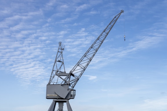 A large metal crane with a long arm extends into a partly cloudy blue sky. The crane has a series of structural supports and a hook hanging from a cable at the end of the arm. The base is stationary and appears sturdy.